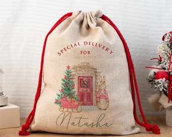 Può includere: Un sacco regalo natalizio beige con chiusura a coulisse rossa. La borsa presenta un'illustrazione ad acquerello di un albero di Natale, una porta rosa, un coniglio e il testo "Special Delivery For Natasha".
