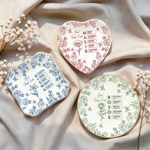 Puede incluir: Tres platos decorativos de cerámica con diseños florales. Uno tiene forma de corazón con flores rojas, otro es cuadrado con flores azules y el tercero es redondo con flores verdes. Cada plato tiene un borde dorado y texto.