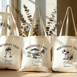 Könnte beinhalten: Drei cremefarbene Canvas-Tragetaschen mit schwarzen Illustrationen und Text. Jede Tasche hat ein anderes Design: Champagnergläser, eine Strandszene und ein Cocktail. Der Text lautet "BIRTHDAY TRIP" sowie ein Name und ein Ort.