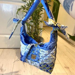 May include: A quilted shoulder bag in shades of blue, white, and green, with a floral design. The bag has a rounded shape with a blue interior and matching blue straps. A small tag is sewn on the front.