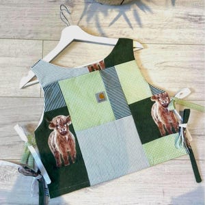 May include: A patchwork sleeveless top featuring a mix of dark green fabric, light green gingham, and thin blue and white stripes. The garment includes two printed images of brown Highland cows and a small Carhartt brand label on the chest.