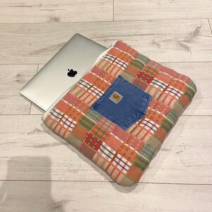 May include: A patchwork laptop sleeve with a large blue denim pocket featuring a Carhartt logo. The sleeve is made of various shades of pink, red, green, and white plaid fabric.