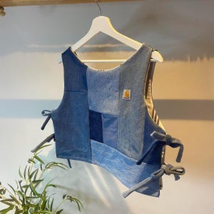 May include: A patchwork denim vest in various shades of blue, hanging on a white hanger. The vest features a mix of denim squares and rectangles, with tie closures on the sides. A small, square Carhartt logo is visible.