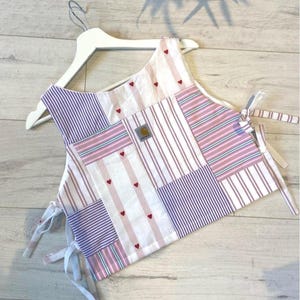 May include: A sleeveless patchwork top featuring a mix of white, pink, and purple striped fabric panels. The garment includes small red heart embroidery, side tie closures, and a rectangular Carhartt brand patch on the front centre panel.