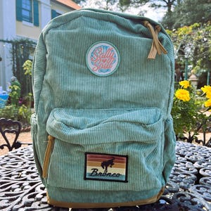 Peut inclure: Un sac à dos en velours côtelé turquoise avec une poche avant et une poignée supérieure. Le sac à dos comporte deux écussons : l'un avec le texte "Salty Soul" et l'autre avec une silhouette de cheval et le mot "Bronco". La bordure inférieure est marron.