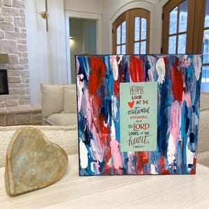 Puede incluir: Pintura abstracta sobre lienzo con una combinación de colores azul, rojo y blanco. La pintura presenta una cita sobre el corazón: "Las personas miran la apariencia exterior, pero el Señor mira el corazón." 1 Samuel 16:7