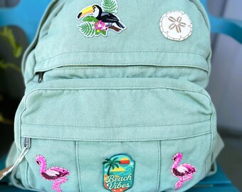 Puede incluir: Una mochila verde claro con múltiples parches. Los parches incluyen un tucán, un dólar de arena, dos flamencos rosas y un parche con una palmera y las palabras "Beach Vibes". La mochila tiene cierre de cremallera.
