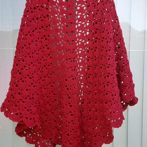 May include: A handmade, red crochet shawl with an openwork design. The shawl is draped over a hanger, showcasing its intricate pattern and scalloped edges. The vibrant red color and delicate texture make it a cozy and stylish accessory.