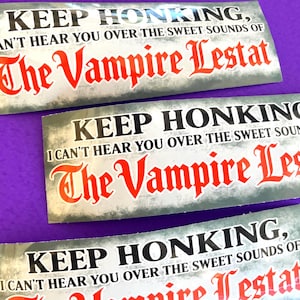 May include: Three rectangular bumper stickers with a distressed grey background. The text reads: KEEP HONKING, I CAN'T HEAR YOU OVER THE SWEET SOUNDS OF The Vampire Lestat. The words are printed in black and bold red gothic-style lettering.
