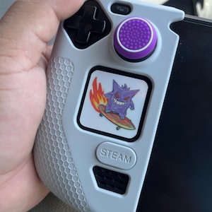 May include: A light grey handheld gaming controller cover featuring a textured honeycomb grip pattern, a black directional pad, and a purple joystick cap. A square sticker displays a purple Gengar character riding a flaming skateboard.