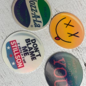 May include: Four round, colorful stickers with various designs and text. One features a green and blue soda logo, another a yellow smiley face with crossed-out eyes, and two with political statements.