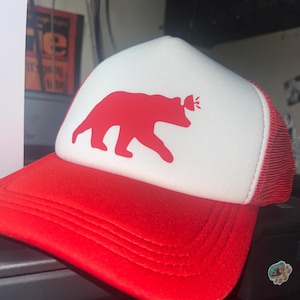 May include: A red and white trucker hat featuring a solid red bear silhouette graphic on the front white foam panel. The hat has a red brim, red mesh back panels, and a small red icon above the bear's head. The design is centered on the front.