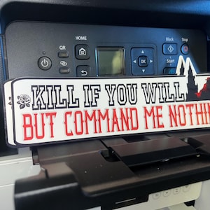 以下が含まれることがあります： 「KILL IF YOU WILL BUT COMMAND ME NOTHING」というテキストが黒と赤で書かれた長方形のサイン。サインにはバラと人物と建物のシルエットがあります。サインはプリンターの上に置かれています。