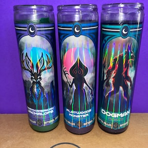 May include: Three tall cylindrical glass candles featuring holographic labels with cryptid illustrations. The labels depict a Jackalope from Wyoming, a Flatwoods Monster from West Virginia, and a Dogman from Wisconsin, each with a crescent moon icon.