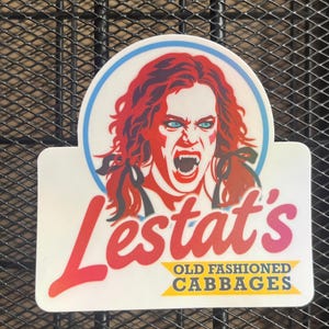 May include: A rectangular sticker with a circular top featuring a red-toned illustration of a person with long hair and fangs. The text reads Lestat's in large red script and OLD FASHIONED CABBAGES in blue letters on a yellow banner background.