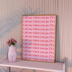 May include: A pink framed print with the text "Call me when you go ZTA" repeated 12 times. The print is hanging on a white wall with a pink ceramic vase with flowers on a white shelf.