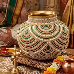 Könnte beinhalten: Ein dekorativer, runder, goldfarbener Topf mit aufwendiger Perlenarbeit in Weiß, Grün und Rot. Der Topf hat einen goldenen Rand und ist mit grünen Edelsteinen verziert. Er steht auf einem Holzständer, mit einer kleinen Messinglampe und Blumen in der Nähe.