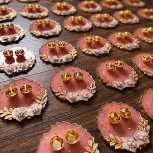 May include: A collection of circular, dusty rose-colored resin ring holders featuring gold-toned metallic leaf accents and white floral embellishments. Each holder contains two small, gold-toned circular cups designed to hold rings securely.