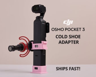 May include: A black DJI Osmo Pocket 3 camera with a pink cold shoe adapter and a red microphone attached to it. The text "OSMO POCKET 3 COLD SHOE ADAPTER" is written in white on a black background. The text "SHIPS FAST!" is written in white on a black background.