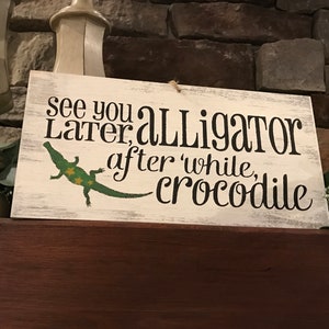 May include: A white wooden sign with black text that reads "See you later, alligator, after a while, crocodile". A green alligator with yellow spots is pictured on the sign.