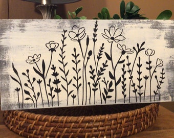 May include: A white wooden sign with a black silhouette of a field of wildflowers. The sign has a distressed, rustic look.