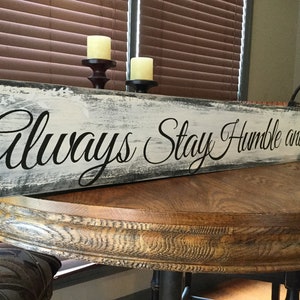 May include: A white wooden sign with black lettering that reads "Always Stay Humble and Kind". The sign has a distressed, weathered look and is sitting on a wooden table.