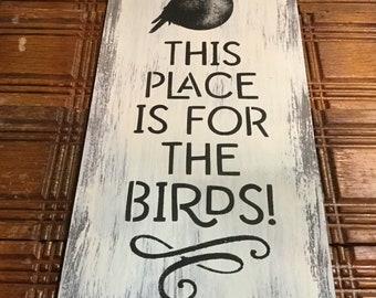 Könnte beinhalten: Ein weißes Holzschild mit schwarzem, bemaltem Text und einer schwarzen Silhouette eines Vogels. Das Schild lautet "This Place Is For The Birds!" mit einer dekorativen Verzierung am unteren Rand.