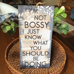 May include: A wooden sign with a gray background and white and gold painted stripes. The sign has white lettering that reads "I'm not bossy I just know what you should be doing".