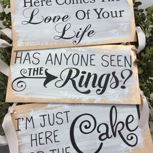 May include: Three wooden wedding signs with white painted backgrounds and gold edges. The signs read: "Here Comes The Love Of Your Life," "Has Anyone Seen The Rings?" and "I'm Just Here For The Cake!" Each sign has a cream-colored ribbon for hanging.