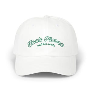 May include: White baseball cap with the embroidered text "Fuck Pierre and his seeds" in green script. The cap has a curved brim and a rounded crown. The text is centered on the front of the cap.