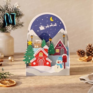Può includere: Un biglietto di carta tridimensionale che raffigura un villaggio invernale con una casa rossa, un pupazzo di neve, pini verdi e lampioni. Lo sfondo mostra un cielo notturno blu scuro con una luna crescente gialla e una slitta volante.