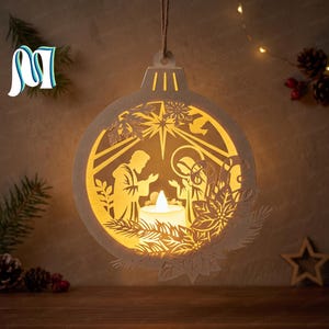 May include: A circular white laser-cut wooden ornament featuring a nativity scene silhouette with a central glowing LED tea light. The design includes a star, foliage, and a hanging twine loop, creating a warm light effect through the cutouts.