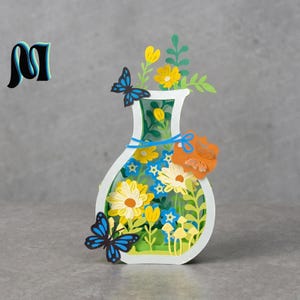 May include: A paper art vase filled with yellow and white flowers, blue butterflies, and green leaves. The vase is white with a green interior and a blue ribbon. The artwork has a 3D effect. The piece is made of layered paper.