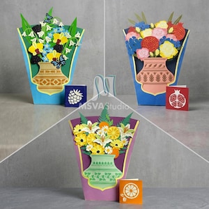 May include: Three pop-up greeting cards featuring paper bouquets in vases. One shows bluebells and butterflies, one shows roses and pomegranates, and one shows citrus slices and daisies. Each card includes a small matching square note card.