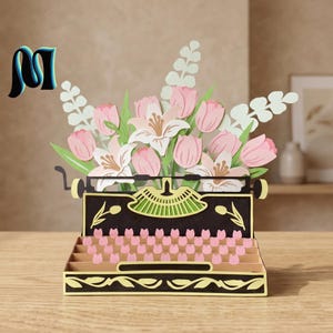 Peut inclure: Une carte pop-up en papier en trois dimensions représentant une machine à écrire noire avec des accents dorés. La machine est remplie d'un bouquet de tulipes roses et de lys blancs, avec des touches en forme de tulipes roses et un logo M.