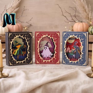 Puede incluir: Tres cajas de sombras de papel rectangulares diseñadas como libros. La caja negra presenta un espantapájaros, la roja muestra un fantasma blanco y la azul una bruja en un cementerio. Cada una tiene un marco decorativo con bordes irregulares.