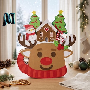 Op de afbeelding: Een meerlaagse papieren decoratie met een beige rendiergezicht met een rode neus en sjaal. De bovenkant toont een peperkoekhuisje, een sneeuwpop, twee groene kerstbomen, een zuurstok en een takje met hulstbladeren en rode bessen.