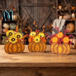 May include: Three decorative pumpkin-shaped centerpieces featuring intricate cut-out designs. The left features sunflowers, the center displays yellow flowers with black bat silhouettes, and the right showcases orange roses and dark feathers.
