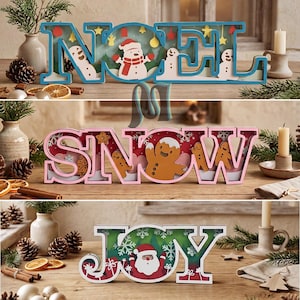 Pode incluir: Três placas decorativas de madeira com letras recortadas que formam as palavras NOEL, SNOW e JOY. A placa NOEL é azul com bonecos de neve, a placa SNOW é rosa com figuras de pão de mel e a placa JOY é branca com uma ilustração do Papai Noel.