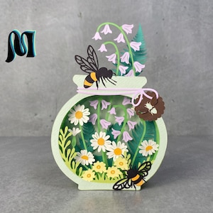 May include: A paper art piece in the shape of a jar, with layered daisies, bellflowers, and bees. The jar is light green with a pink ribbon and a brown flower accent. The bees have black and yellow stripes. The background is a light grey.