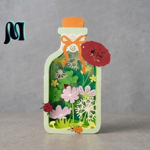 Puede incluir: Una pieza de arte en papel en capas con forma de botella, que presenta una escena de jardín con flores, tréboles y mariquitas. La botella tiene un marco verde claro, un corcho naranja y una etiqueta roja con un recorte de mariquita.
