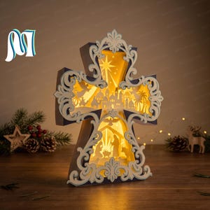 May include: A cross-shaped light box featuring a layered paper cut-out design. The top section displays a star over a city silhouette, while the bottom section depicts a nativity scene with figures, a dove, and animals, all illuminated by a warm glow.