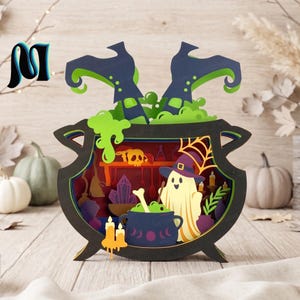 May include: A multi-layered paper craft display shaped like a black cauldron. It features a ghost wearing a purple hat, a smaller cauldron with green liquid, a skull, crystals, and candles. Two witch boots in black and green emerge from the top.