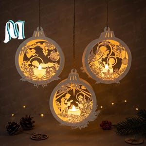 May include: Three white, circular, laser-cut wooden ornaments hanging by strings. Each ornament features a different intricate nativity scene silhouette illuminated from within by a small, warm-glowing LED tea light candle.