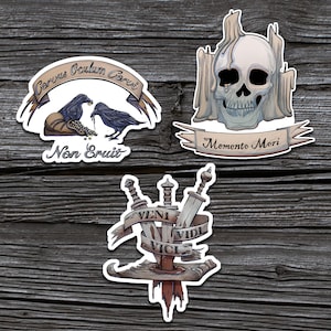 May include: Three vintage-style stickers with Latin phrases and imagery. The first sticker features two black ravens with a bag of gold coins and the text "Corvus Oculum Corvi Non Erit". The second sticker features a skull with candles and the text "Memento Mori". The third sticker features three swords with the text "Veni Vidi Vici".