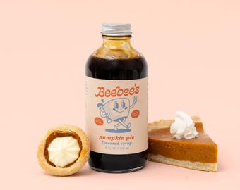 Könnte beinhalten: Eine Glasflasche mit Beebee's Kürbiskuchen-Sirup mit silbernem Verschluss, ein Etikett mit einer Cartoon-Tassenfigur, daneben ein kleines rundes Gebäck mit weißer Creme und ein Stück Kürbiskuchen mit einem Klecks Schlagsahne darauf.