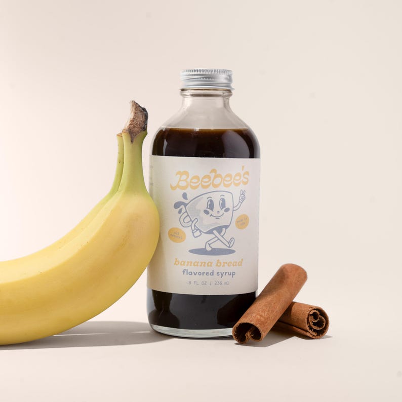 May include: A glass bottle of Beebee's banana bread flavored syrup with a cartoon illustration of a mug on the label. A banana and two cinnamon sticks are in the foreground.