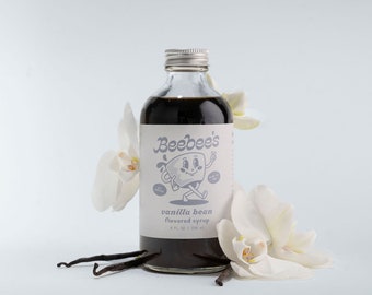 May include: A glass bottle of vanilla bean flavored syrup with a white label that says "Beebee's" and features a cartoon illustration of a mug with a face. The bottle is surrounded by white orchid flowers and vanilla beans.