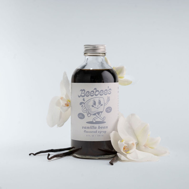 May include: A glass bottle of vanilla bean flavored syrup with a white label that says "Beebee's" and features a cartoon illustration of a mug with a face. The bottle is surrounded by white orchid flowers and vanilla beans.