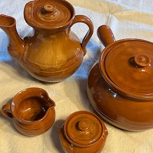 May include: A set of four brown glazed ceramic kitchen items including a teapot with a lid, a large lidded pot with a side handle, a small creamer pitcher, and a small lidded sugar bowl. Each piece features a smooth, glossy finish.